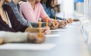 Cropped view of a multiracial group of young men and women sitting in a row at a table, writing with pencils on paper.  They are taking a test or filling out an application.  Focus is on the hand of the young man in the middle in the gray shirt.