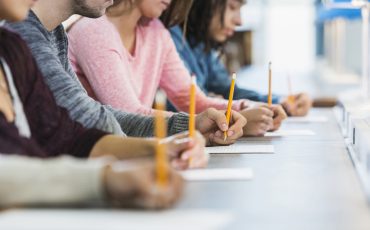 Cropped view of a multiracial group of young men and women sitting in a row at a table, writing with pencils on paper.  They are taking a test or filling out an application.  Focus is on the hand of the young man in the middle in the gray shirt.