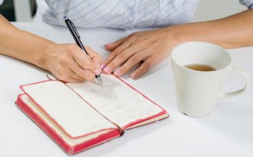 the-hand-of-a-woman-with-a-pen-writes-in-her-diary-next-on-the-table-is-a-cup-of-tea